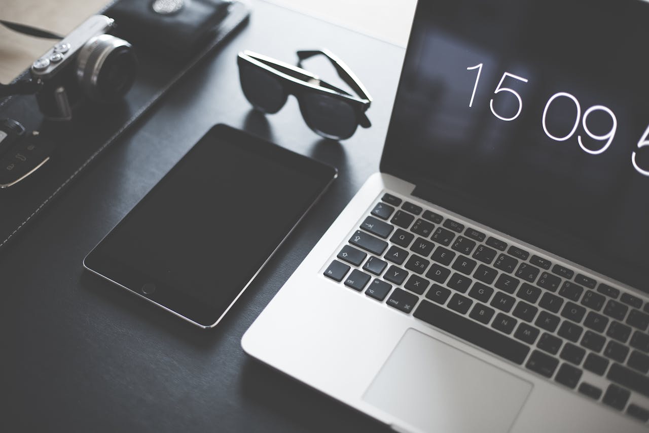 digital A flat lay of a modern workspace featuring a laptop, tablet, camera, and sunglasses.
