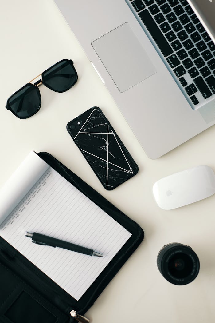 A clean workspace featuring a laptop, smartphone, sunglasses, and stationery on a white surface.